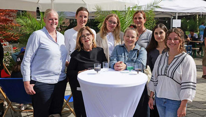Mehrere Mitarbeiterinnen stehen um einen weißen Stehtisch und blicken fröhlich in die Kamera beim Albertinen-Sommerfest für die Mitarbeitenden aus den unterschiedlichen Einrichtungen der Immanuel Albertinen Diakonie am Standort Schnelsen auf dem Platz vor der Kirche am Albertinen Krankenhaus.