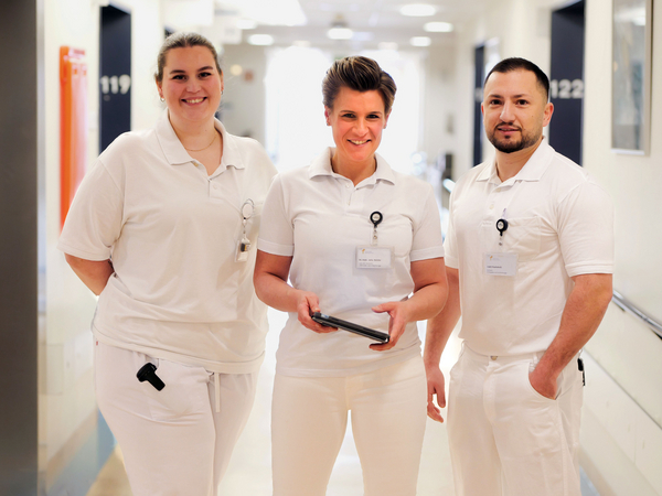 Physician Assistant Hanna Lübbe, Zentrumsleiterin Dr. med. Julia M. Reinke mit Tablet-Computer in der Hand und Facharzt Irakli Paatishvili stehen nebeneinander in weißer Funktionskleidung im Stationsflur und blicken in die Kamera im auf Hüftprothesen, Knieprothesen und Schulterprothesen spezialisierten Albertinen Endoprothetikzentrum Hamburg