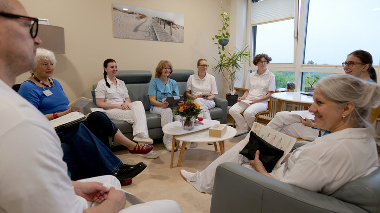 Ein interdisziplinäre Team sitzt überwiegend in Funktionskleidung zu einer Besprechung in einem hell und modern eingerichteten Raum der Klinik für Innere Medizin mit Schwerpunkt Palliativmedizin im Albertinen Krankenhaus in Hamburg.