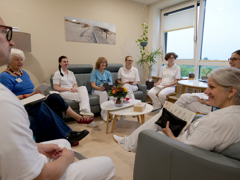 Ein interdisziplinäre Team sitzt überwiegend in Funktionskleidung zu einer Besprechung in einem hell und modern eingerichteten Raum der Klinik für Innere Medizin mit Schwerpunkt Palliativmedizin im Albertinen Krankenhaus in Hamburg.