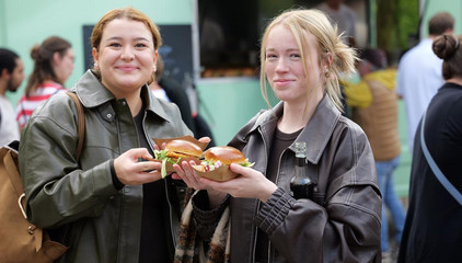 Zwei Besucherinnen mit frischen Burgern in den Händen blicken in die Kamera beim Albertinen-Sommerfest für die Mitarbeitenden aus den unterschiedlichen Einrichtungen der Immanuel Albertinen Diakonie am Standort Schnelsen auf dem Platz vor der Kirche am Albertinen Krankenhaus.