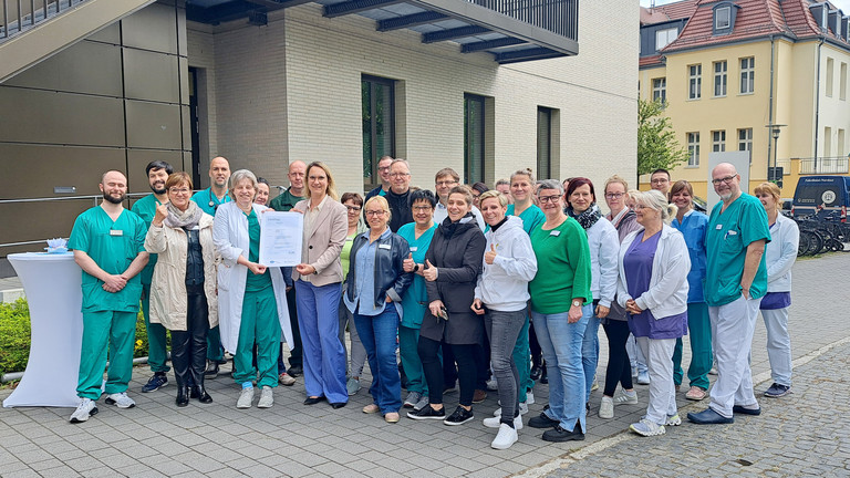 Mitarbeitende des Immanuel Klinikums Bernau vor dem Klinikgebäude mit dem Zertifikat Qualitätszertifikat DIN EN ISO 9001:2015 