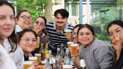 Mehrere Besucherinnen und Besucher prosten mit Bierbechern in den Händen fröhlich in die Kamera beim Albertinen-Sommerfest für die Mitarbeitenden aus den unterschiedlichen Einrichtungen der Immanuel Albertinen Diakonie am Standort Schnelsen auf dem Platz vor der Kirche am Albertinen Krankenhaus.