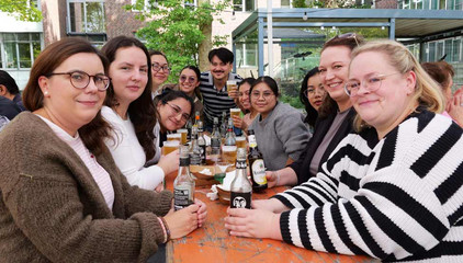 Eine Gruppe fröhlicher Besucher sitzt auf einer Bierbank mit Getränken und Essen beim Albertinen-Sommerfest für die Mitarbeitenden aus den unterschiedlichen Einrichtungen der Immanuel Albertinen Diakonie am Standort Schnelsen auf dem Platz vor der Kirche am Albertinen Krankenhaus.