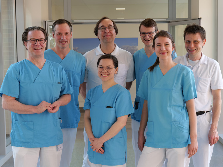 Das Team Des Mitralklappen-Zentrums des Albertinen Herz- und Gefäßzentrums am Standort Hamburg Schnelsen lächelt auf einem Flur in Arztkleidung in die Kamera
