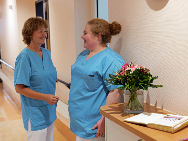 Zwei Pflegekräfte der Palliativpflege unterhalten sich in blauer Funktionskleidung auf einem hellen Stationsflur neben einem mit Blumen geschmückten Stationstisch in der Klinik für Innere Medizin mit Schwerpunkt Palliativmedizin im Albertinen Krankenhaus in Hamburg. 