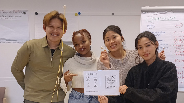 Vier Auszubildende präsentieren im Klassenraum das Ergebnis einer Teamaufgabe: einen freistehenden Turm aus Spaghetti und Marshmallows auf einem Tisch.