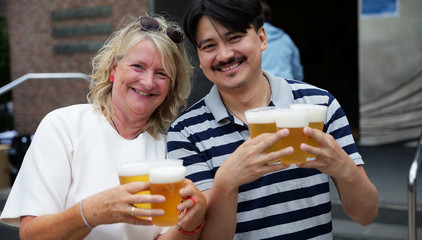 Eine Mitarbeiterin und ein Mitarbeiter halten mehrere Bierbecher in die Kamera beim Albertinen-Sommerfest für die Mitarbeitenden aus den unterschiedlichen Einrichtungen der Immanuel Albertinen Diakonie am Standort Schnelsen auf dem Platz vor der Kirche am Albertinen Krankenhaus.