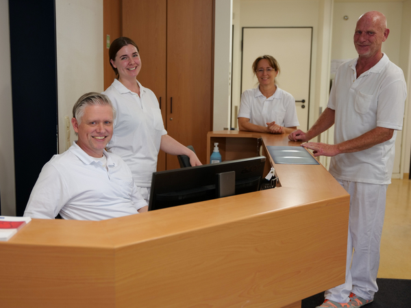 Mehrere Therapeutinnen und Therapeuten in weißer Funktionskleidung stehen am Empfangstresen bei Physikofit für Physiotherapie im Interdisziplinären Therapiezentrum im Albertinen Krankenhaus in Hamburg und blicken in die Kamera