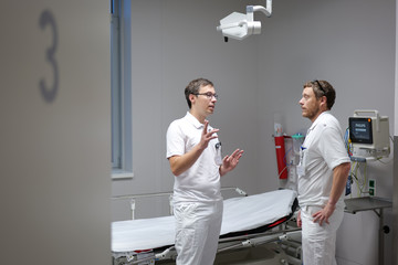 Zwei Ärzte unterhalten sich in einem Behandlungsraum der Zentralen Notaufnahme neben einer Untersuchungsliege und einem Patientenmonitor.