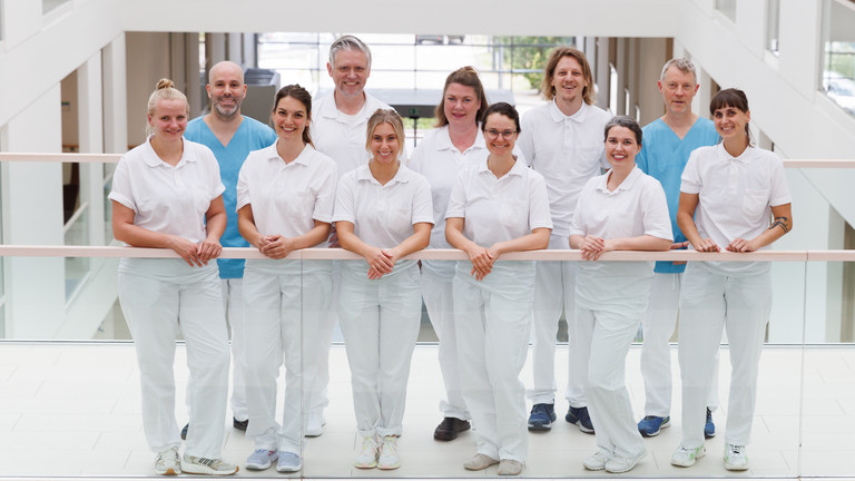 Das Team der Neurologie des Interdisziplinären Therapiezentrums im Albertinenkrankenhaus steht in Dienstkleidung freundlich in die Kamera blickend auf dem Flur
