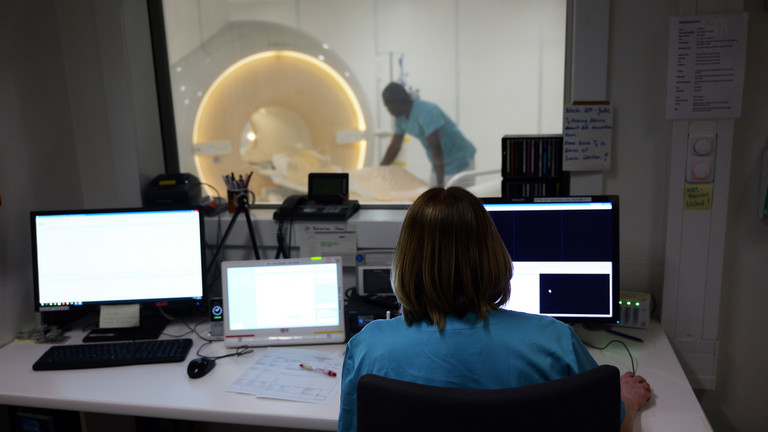 Blick über die Schulter einer Mitarbeiterin an einem PC im Vorraum eines MRTs, im Hintergrund sieht man einen Mitarbeiter am Gerät