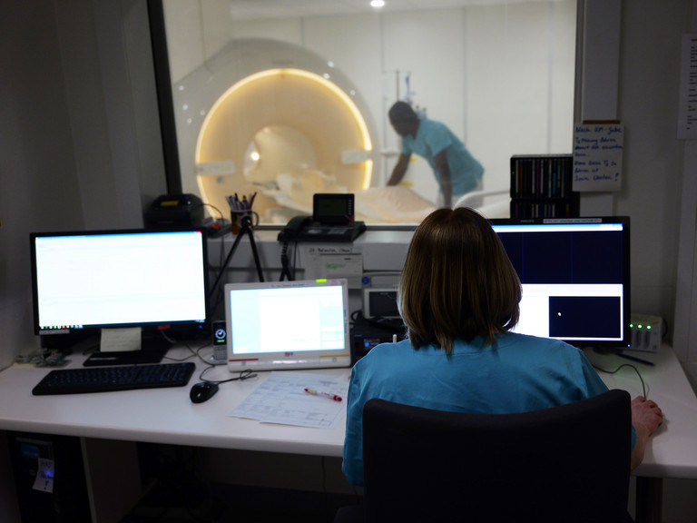 Blick über die Schulter einer Mitarbeiterin an einem PC im Vorraum eines MRTs, im Hintergrund sieht man einen Mitarbeiter am Gerät