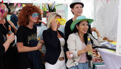 Ein Gruppe Besucher mit bunten Sonnenbrillen und farbigen Hüten steht zusammen beim Albertinen-Sommerfest für die Mitarbeitenden aus den unterschiedlichen Einrichtungen der Immanuel Albertinen Diakonie am Standort Schnelsen auf dem Platz vor der Kirche am Albertinen Krankenhaus.