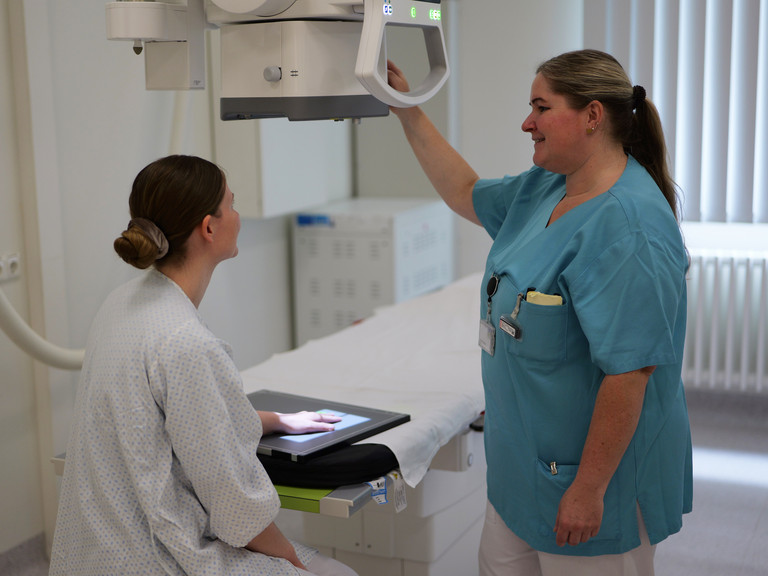 Eine Patientin bekommt ein digitales Röntgen der Hand, eine Mitarbeiterin in blauem Kasak bedient das Gerät