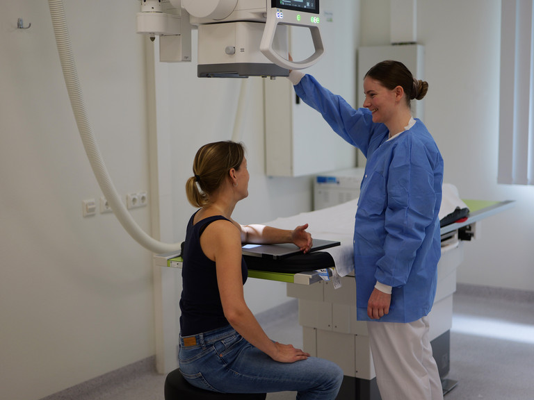 Eine Frau erhält ein Röntgen von ihrem Ellenbogen, eine Mitarbeiterin in Arbeitskleidung bedient das digitale Röntgen