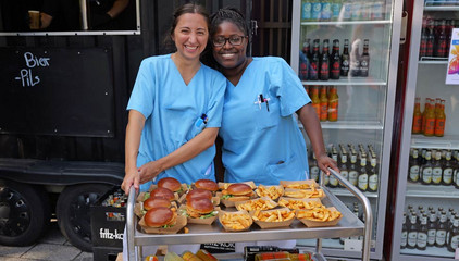 Zwei Mitarbeiterinnen in blauer Funktionskleidung stehen hinter einem Servierwagen mit frischen Burgern und Pommes und blicken fröhlich in die Kamera beim Albertinen-Sommerfest für die Mitarbeitenden aus den unterschiedlichen Einrichtungen der Immanuel Albertinen Diakonie am Standort Schnelsen auf dem Platz vor der Kirche am Albertinen Krankenhaus.