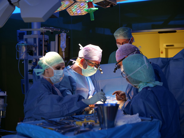 Prof. Dr. Hanke steht mit dem OP-Team im tiefblau erleuchteten Operationssaal und blickt mithilfe einer LED-Stirnlampe auf das Operationsfeld während einer Herz-Operation im Albertinen Herz- und Gefäßzentrum in Hamburg