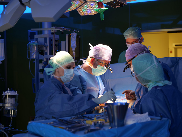 Prof. Dr. Hanke steht mit dem OP-Team im tiefblau erleuchteten Operationssaal und blickt mithilfe einer LED-Stirnlampe auf das Operationsfeld während einer Herz-Operation im Albertinen Herz- und Gefäßzentrum in Hamburg